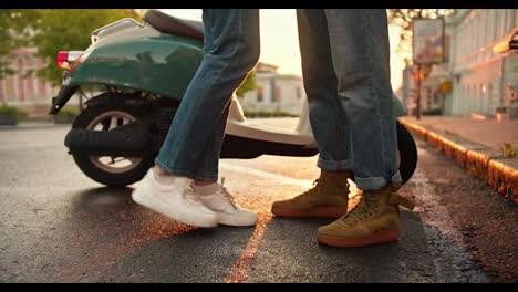 Close-up-shot-of-a-girl-standing-on-her-toes-to-hug-her-boyfriend-near-their-moped-on-the-street-during-dawn-in-summer.-Legs-of-a-guy-and-a-girl-in-denim-pants-standing-opposite-each-other-against-of-a-bright-sunny-dawn-in-summer