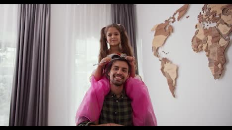 Portrait-of-a-happy-family-little-brunette-girl-in-a-pink-fairy-costume-with-a-magic-wand-sitting-on-the-shoulders-of-her-Happy-brunette-male-father-in-a-Green-checkered-shirt-in-a-modern-home