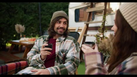 Over-shoulder:-A-happy-brunette-guy-with-a-green-hat-in-a-checkered-shirt-and-his-brunette-girlfriend-drink-tea-from-special-thermos-cups-during-their-picnic-outside-the-city-with-a-trailer