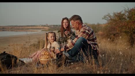 Un-Hombre-Moreno-Feliz-Con-Cabello-Gris-Con-Una-Camisa-A-Cuadros-Se-Sienta-En-El-Suelo-Sobre-Una-Colchoneta-Especial-Durante-Un-Picnic-Y-Bebe-Té-De-Un-Termo-Con-Su-Esposa-Y-Su-Pequeña-Hija-Durante-Un-Picnic-Fuera-De-La-Ciudad-Cerca-De-Un-Estanque-En-Verano.