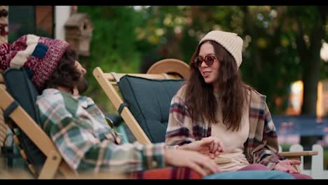 Happy-brunette-girl-in-a-white-hat-with-round-glasses-in-a-plaid-shirt-communicates-with-her-brunette-boyfriend-on-dark-green-chairs-near-a-trailer-in-the-camp-during-her-picnic-outside-the-city-in-the-summer