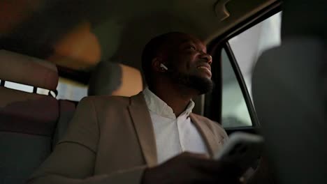 A-happy-man-with-Black-skin-with-a-beard-in-a-brown-suit-sits-on-the-passenger-seat-and-listens-to-music-in-white-wireless-headphones-during-his-vacation-in-a-modern-car-interior-in-the-city