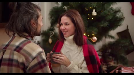 Close-up-shot-of-a-guy-and-a-brunette-girl-in-a-checkered-shirt-and-a-red-blanket-sitting-talking-and-looking-at-each-other,-a-girl-drinking-a-hot-drink-from-a-gray-glass-near-a-green-decorated-Christmas-tree-in-the-winter-evening