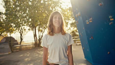 Portrait-of-a-happy-blonde-rock-climber-girl-in-a-white-T-shirt-posing-and-looking-at-the-camera-while-near-a-blue-climbing-wall-on-a-sunny-day-in-summer