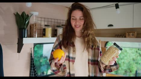 A-happy-brunette-girl-in-a-plaid-shirt-holds-an-orange-and-a-jar-of-pasta-in-her-hands-and-chooses-something-from-them-during-her-vacation-in-a-trailer-in-a-camp-in-the-city-in-the-summer