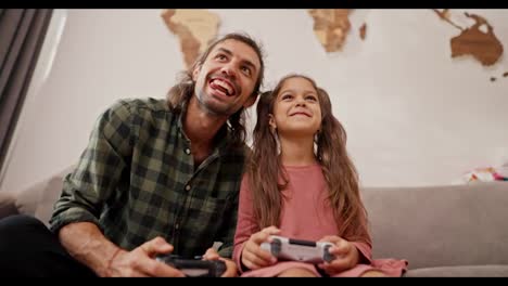 A-little-brunette-girl-with-a-ponytail-hairstyle-in-a-pink-dress-plays-with-a-joystick-together-with-her-happy-father,-a-brunette-man-in-a-Green-checkered-shirt-sitting-on-a-gray-sofa-in-a-modern-apartment