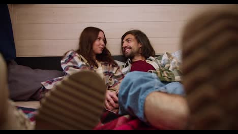 Close-up-shot-of-a-brunette-girl-in-a-plaid-shirt-lying-on-a-bed-in-a-trailer-with-her-brunette-boyfriend-and-they-put-their-legs-forward-touching.-Happy-couple-guy-and-girl-brunettes-in-plaid-shirts-relax-in-a-trailer-outside-the-city-during-a-picnic-at-a-camp-in-the-summer