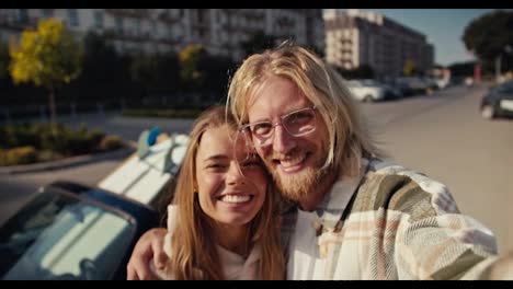 Portrait-of-a-happy-couple,-a-blond-guy-in-glasses-with-a-beard-takes-a-selfie-with-his-blonde-girlfriend-who-smiles-widely-in-a-White-hoodie.-They-show-the-V-sign-and-have-fun-in-the-city