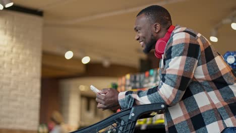 A-happy-and-joyful-man-with-Black-skin-and-a-short-haircut-and-beard-in-a-plaid-shirt-and-wireless-headphones-looks-at-the-list-of-products-on-his-phone-that-he-needs-to-buy-while-going-to-the-supermarket-with-a-cart