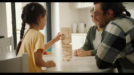 Rear-view-of-a-little-brunette-girl-in-a-yellow-dress-playing-Jenga-with-her-parents-in-a-white-kitchen-in-a-modern-apartment.-A-little-girl-plays-a-game-for-developing-hand-motor-skills-with-her-parents-and-communicates-with-them-while-sitting-in-the-kitchen-at-home