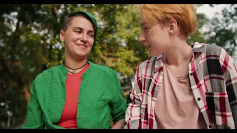 Close-up-shot-of-a-girl-with-short-green-hair-in-a-green-shirt-and-a-blonde-girl-with-short-hair-in-a-checkered-shirt-talking-on-their-date-in-the-park.-Two-lesbians-in-the-park-on-a-date