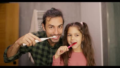 Portrait-of-a-brunette-man-in-a-green-checkered-shirt-brushing-his-teeth-with-his-little-daughter-brunette-girl-in-a-pink-dress.-They-look-in-the-mirror,-look-at-the-camera-and-brush-their-teeth-in-a-modern-bathroom