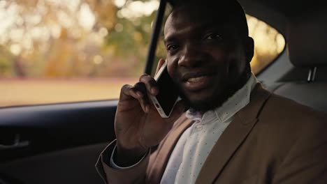 Portrait-of-a-happy-Black-skinned-businessman-in-a-brown-jacket-who-communicates-on-the-phone-while-driving-in-the-passenger-seat-in-a-modern-car-outside-the-city-during-a-business-trip