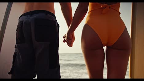 Close-up-shot-of-a-guy-in-blue-shorts-and-a-girl-in-an-orange-swimsuit-standing-next-to-each-other-holding-surfboards-and-holding-hands-looking-towards-the-sea-in-the-morning