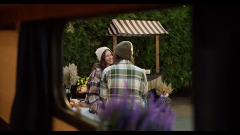 Shooting-from-the-window-of-a-trailer,-a-brunette-girl-in-a-white-hat-and-a-pink-checkered-shirt-communicates-with-her-brunette-boyfriend-in-a-checkered-shirt-who-is-sitting-at-a-table-outside-the-city-in-a-camp-during-their-picnic-in-the-summer