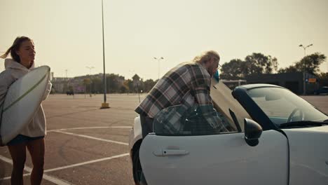 Ein-Blonder-Typ-Mit-Brille-Und-Kariertem-Hemd-Packt-Sein-Surfbrett-In-Ein-Weißes-Cabrio-Und-Auch-Das-Brett-Seiner-Blonden-Freundin.-Glückliches-Paar-Macht-Sich-Bereit,-Nach-Dem-Surfen-In-Der-Stadt-Nach-Hause-Zu-Fahren