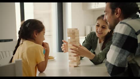 Happy-young-family-brunette-girl-in-a-green-jacket,-her-husband,-brunette-guy-in-a-checkered-shirt-and-their-little-daughter,-brunette-girl-in-a-yellow-t-shirt,-play-Jenga,-a-game-for-developing-finger-motor-skills,-in-a-modern-kitchen-in-an-apartment