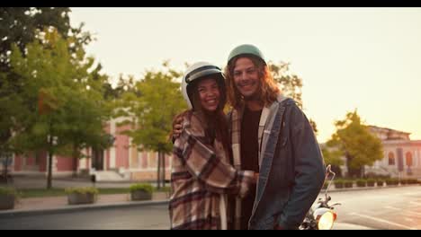 Portrait-of-a-happy-guy-with-curly-hair-in-a-helmet-and-a-denim-shirt-stands-next-to-and-hugs-his-brunette-girlfriend-with-curly-hair-in-a-plaid-shirt-and-a-white-helmet-near-his-moped-on-a-wide-street