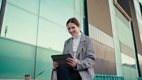 A-happy-brunette-girl-with-round-glasses-in-a-striped-gray-jacket-sits-near-a-large-building-and-works-on-her-black-tablet-with-a-mug-of-coffee-during-the-day-in-the-city