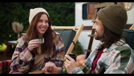 Close-up-shot-of-a-happy-brunette-girl-drinking-tea-from-a-thermos-mug-together-with-her-brunette-boyfriend-in-a-green-checkered-shirt-who-holds-her-hand-and-communicates-with-her-during-his-picnic-outside-the-city-near-a-trailer-on-chairs