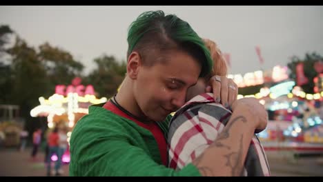 Close-up-shot-of-a-girl-with-short-green-hair-in-a-checkered-green-shirt-and-a-blonde-girl-with-a-short-haircut-in-a-checkered-pink-shirt-hugging-and-looking-at-each-other-during-their-date-in-the-amusement-park