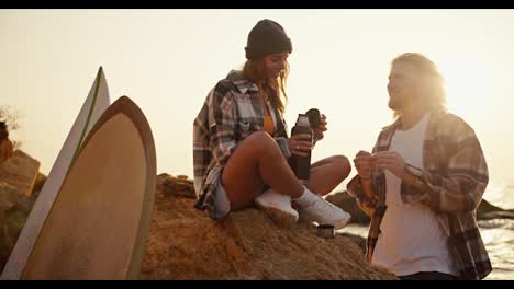 A-blonde-girl-in-a-black-hat-and-a-plaid-shirt-sits-on-a-large-rock-near-the-sea-and-opens-a-thermos-of-tea,-and-her-blonde-boyfriend-looks-at-her.-Happy-couple-guy-and-girl-relaxing-on-a-rocky-beach-near-the-sea-near-their-surfboards-in-the-morning-at-Sunrise