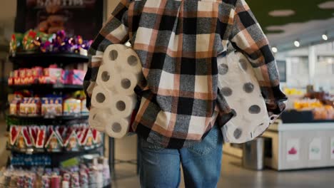 Rear-view-of-a-man-in-a-plaid-shirt-and-blue-jeans-carrying-two-large-packages-of-white-disposable-towels-while-walking-near-the-shelves-in-a-modern-grocery-store