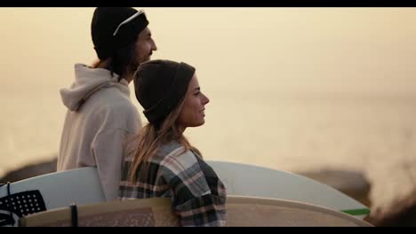 Happy-guy-in-a-white-sweatshirt-and-a-blonde-girl-in-a-checkered-shirt-holding-surfboards-and-looking-towards-the-sea-on-the-earthen-shore-in-the-morning.-A-blonde-girl-in-a-checkered-shirt-and-a-hat-stands-near-her-boyfriend-and-shows-a-sign-of-a-wave-in-the-sea-with-her-hand