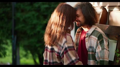 Close-up-shot-of-a-happy-brunette-guy-in-a-Green-checkered-shirt-hugging-his-brunette-girlfriend-in-a-checkered-shirt-near-green-coniferous-trees-Near-his-trailer-in-the-camp-during-a-picnic-in-the-summer