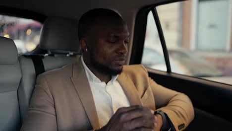 Confident-man-Businessman-with-Black-skin-and-a-beard-in-a-brown-suit-rides-in-a-modern-car-looks-at-his-watch-and-then-out-the-window.-Successful-businessman-driving-in-a-car-to-an-appointed-place