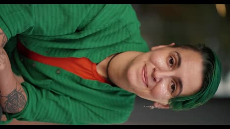 Vertical-video:-portrait-of-a-happy-lesbian-girl-with-a-short-green-haircut-and-in-a-green-shirt-with-tattoos-who-looks-at-the-camera-and-smiles