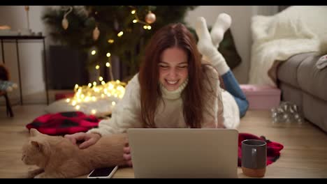 Happy-brunette-girl-in-a-white-sweater-lies-on-the-floor-on-a-red-plaid-watching-a-movie-on-a-laptop-and-stroking-her-cream-colored-cat-in-a-cozy-room-decorated-in-Christmas-style