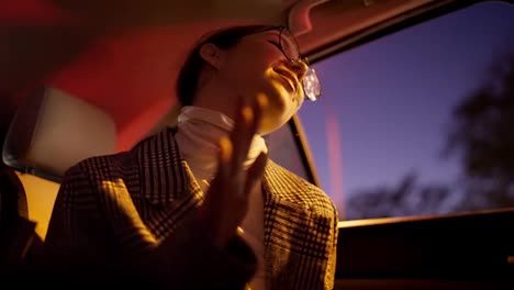 A-happy-brunette-girl-in-round-glasses-in-a-striped-white-and-black-coat-dances-after-a-happy-business-meeting-in-the-cabin-of-a-modern-car-during-a-business-trip-in-the-night-city