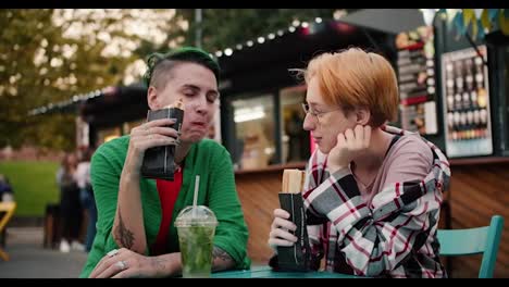 Close-up-shot-of-a-blonde-girl-with-a-short-haircut-and-a-green-short-haired-girl-in-a-Green-shirt-sitting-at-a-table-in-a-street-cafe-eating-walkers-and-drinking-a-cocktail-during-their-date-in-the-summer