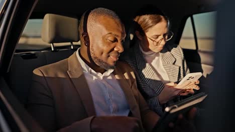 A-confident-man-with-Black-skin-and-a-beard-a-businessman-in-a-brown-suit-explains-his-plans-and-proposals-to-a-businesswoman-in-round-brunette-glasses-using-his-tablet-during-a-general-trip-in-the-cabin-of-a-modern-car