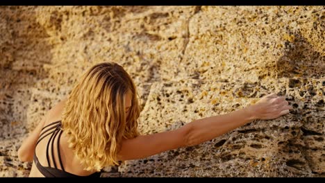 A-blonde-girl-with-curly-hair-in-a-black-top-climbs-up-a-yellow-rock-and-is-looking-for-the-right-place-to-place-her-hand-in-order-to-cling-to-the-surface.-Blonde-girl-climber-climbs-up-a-yellow-rock