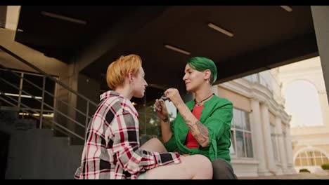 Two-lesbian-girls-with-short-bright-haircuts-in-plaid-shirts-are-sitting-near-a-large-building.-Lesbian-girl-with-green-short-hair-in-a-green-shirt-takes-off-her-glasses-and-hugs-her-blonde-girl-with-short-hair-in-a-plaid-shirt-on-a-date-in-the-summer