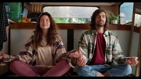 Portrait-of-a-meditating-couple,-a-brunette-girl-in-a-plaid-shirt-and-pink-pants-along-with-her-boyfriend-in-a-Green-plaid-shirt-and-a-red-T-shirt-sitting-in-the-lotus-position-and-meditating-in-their-country-house-or-trailer-during-their-picnic-outside-the-city-in-the-summer