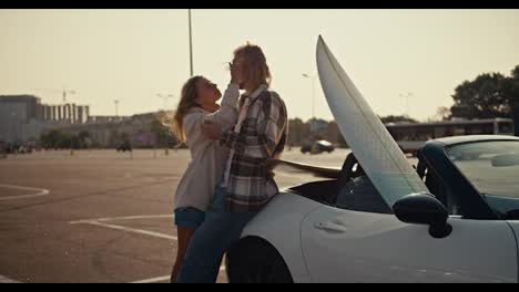 A-blonde-girl-in-a-white-sweatshirt-leans-on-her-blond-boyfriend-in-a-checkered-shirt-and-jeans-who-sits-on-a-white-convertible-and-hugs-his-girlfriend.-Happy-couple,-a-guy-and-a-girl-are-standing-near-a-white-convertible-and-hugging-on-a-square-in-the-city-in-the-morning