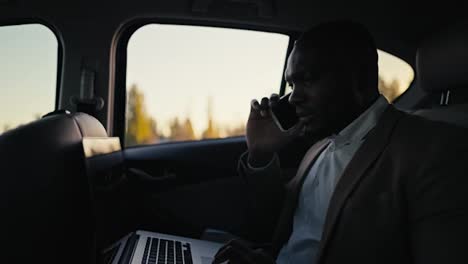 Confident-man-Businessman-with-Black-skin-color-with-a-beard-in-a-brown-jacket-communicates-on-a-smartphone-and-works-on-a-laptop-while-driving-in-the-passenger-seat-in-a-modern-car-outside-the-city