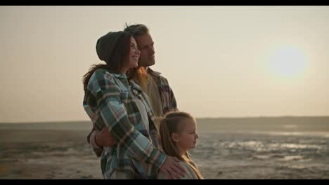 Happy-brunette-woman-in-a-green-checkered-shirt-shows-with-her-hand,-stands-and-hugs-next-to-her-husband,-a-middle-aged-man,-and-a-little-blonde-girl-during-her-vacation-outside-the-city-on-a-deserted-seashore-in-a-summer-evening