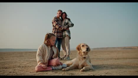 Portrait-of-a-happy-little-blonde-girl-in-a-white-jacket-and-pink-pants-who-sits-and-relaxes-and-pets-her-large-cream-colored-dog-behind-her-are-her-parents-who-are-hugging-and-watching-with-their-little-girl-while-relaxing-on-a-deserted-seashore-in-summer