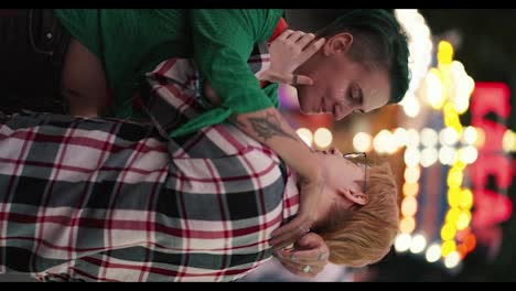 Vertical-video:-A-blonde-girl-with-short-hair-in-a-plaid-shirt-and-a-girl-with-green-hair-with-short-hair-in-a-Green-shirt-hug-during-their-LGBT-date-in-a-bright-amusement-park