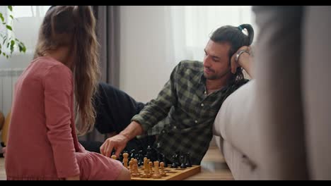 Side-view-of-a-brunette-man-with-stubble-in-a-checkered-shirt-playing-chess-with-his-little-daughter,-a-brunette-girl-in-a-pink-dress,-sitting-on-the-floor-and-leaning-on-a-gray-sofa-in-a-modern-apartment