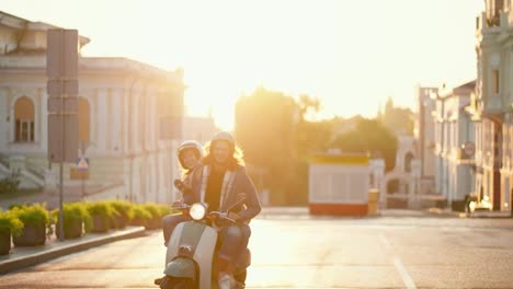 Ein-Glückliches-Paar-Mit-Langen-Lockigen-Haaren,-Einem-Grünen-Motorradhelm-Und-Einer-Jeansjacke-Fährt-Mit-Seiner-Brünetten-Freundin,-Die-Ihm-Hinter-Der-Schulter-Zusieht,-Während-Sie-Auf-Einem-Grünen-Moped-Bei-Einem-Goldenen-Sonnenaufgang-Im-Sommer-Eine-Schöne-Sommerstadtstraße-Entlang-Fährt.