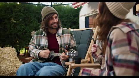 A-happy-brunette-guy-in-a-Green-hat-in-a-Green-checkered-shirt-plays-a-black-ukulele-and-his-brunette-girlfriend-in-a-pink-checkered-shirt-is-a-trailer-dancer-during-their-picnic-in-a-camp-outside-the-city-in-the-summer