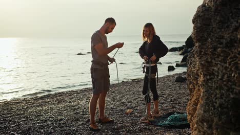 A-blonde-girl-with-curly-hair-in-black-clothes-and-a-brunette-guy-in-a-gray-T-shirt-and-gray-pants-tie-special-knots-on-their-belay-using-special-carabiners-in-order-to-start-rock-climbing-on-a-rocky-seashore-near-the-rocks