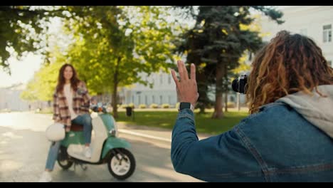 A-guy-with-curly-long-hair-in-a-checkered-jacket-photographs-his-brunette-girlfriend-in-a-checkered-shirt-who-sits-on-a-green-moped-and-shows-her-how-to-pose-correctly-in-a-summer-city-park