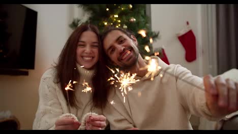 Portrait:-a-happy-couple,-a-guy-and-a-brunette-girl-in-white-sweaters,-looks-at-the-camera-and-smiles-and-holds-sparklers-in-their-hands-that-glow-brightly-and-burn-in-the-New-Year's-atmosphere-near-the-New-Year's-tree-in-a-cozy-home-in-winter