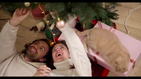 Close-up-shot,-a-guy-and-a-brunette-girl-in-a-white-sweater-are-lying-on-the-floor-on-a-red-blanket-and-looking-at-a-green-decorated-Christmas-tree-near-their-cream-colored-cat-in-a-cozy-room